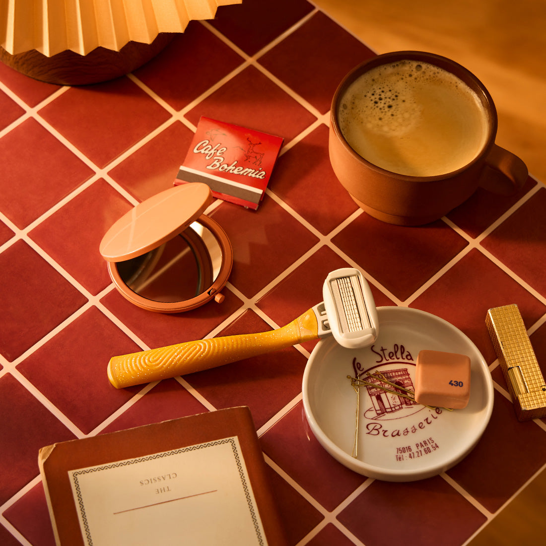 Gold lead razor handle on a tile table with a cup of coffee, book, compact mirror and lipstick.