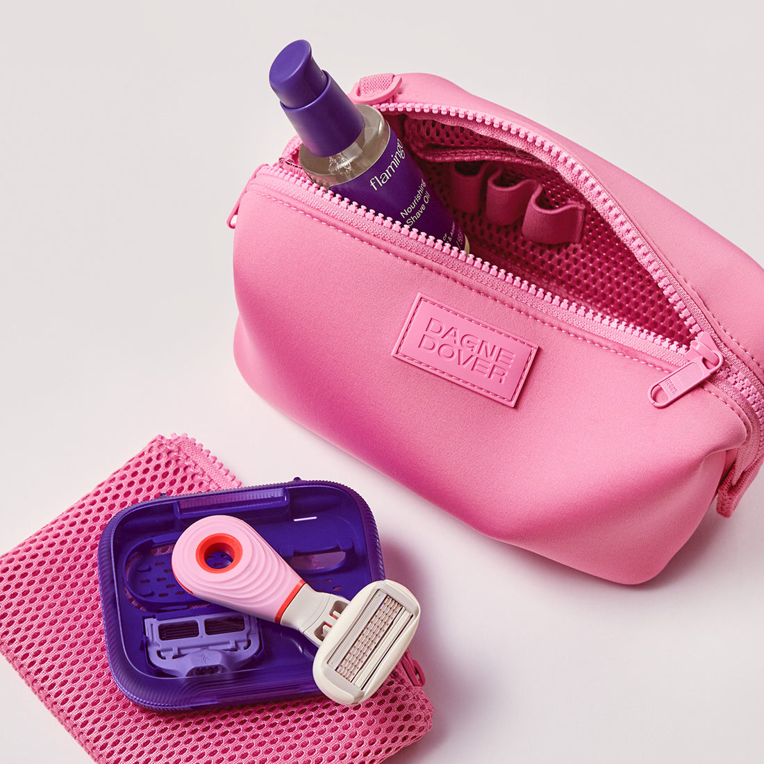Mini Travel Razor on top of a mesh pouch next to the pink Dagne Dover toiletry bag with Shave Oil unzipped on a white background.