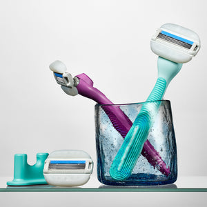 Moisture plus razors in the colors Fig and Seafoam in a glass cup, next to one Moisture Plus blade and the Sea foam shower holder.