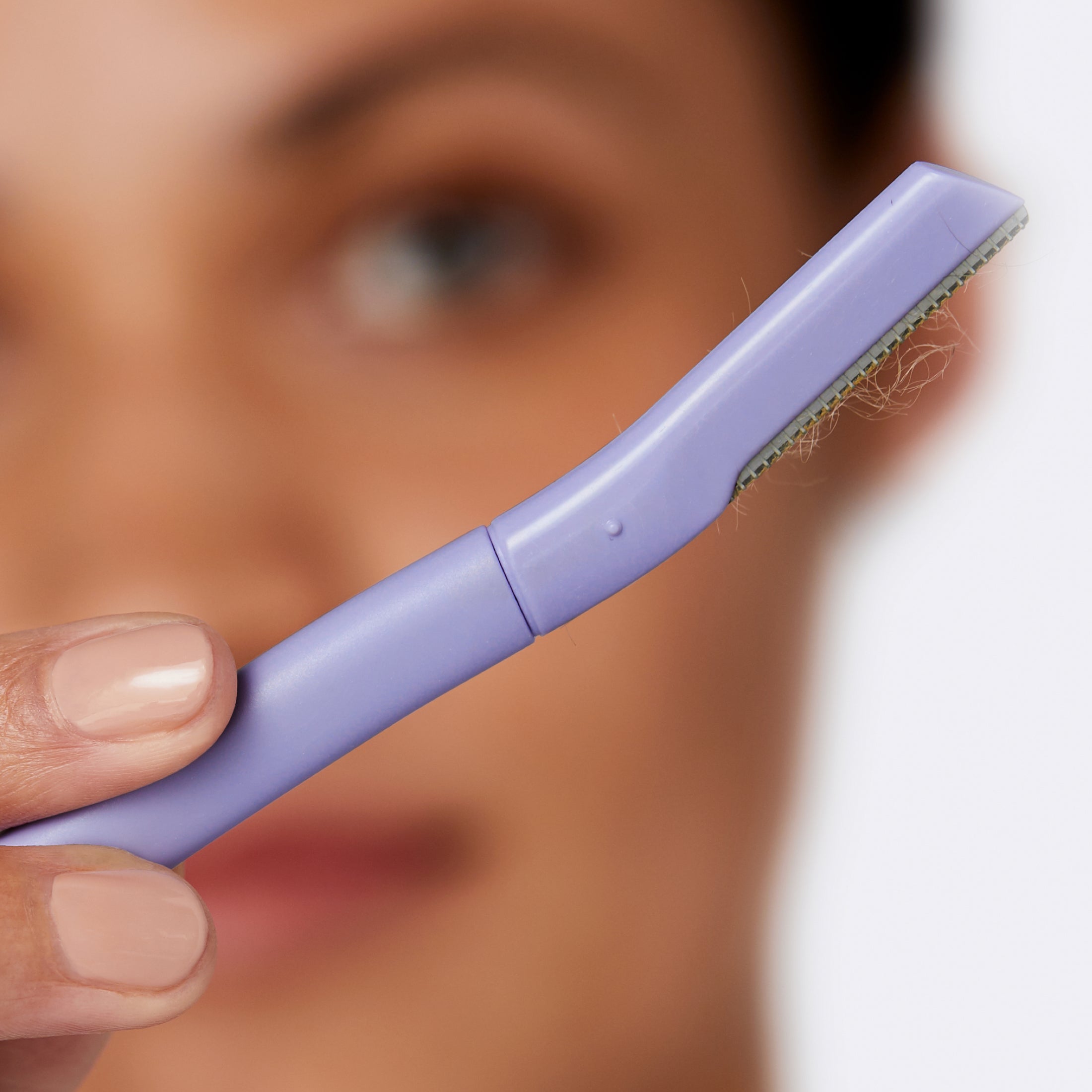 Close up of a woman holding a lilac Flamingo Disposable Dermaplane Razor with peach fuzz on it that she's removed from her face