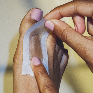 Closeup of a woman applying a small Flamingo Wax strip to her big toe, her nails and toenails are painted a light pink color