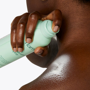 Woman holding Flamingo Light Hydrating spray over her shoulder and spraying her back with the light formula