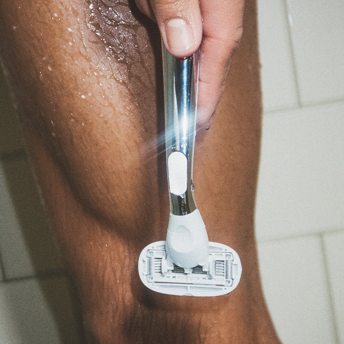 Closeup of a person holding a Flamingo Polished Chrome Razor in front of their leg in the bathroom