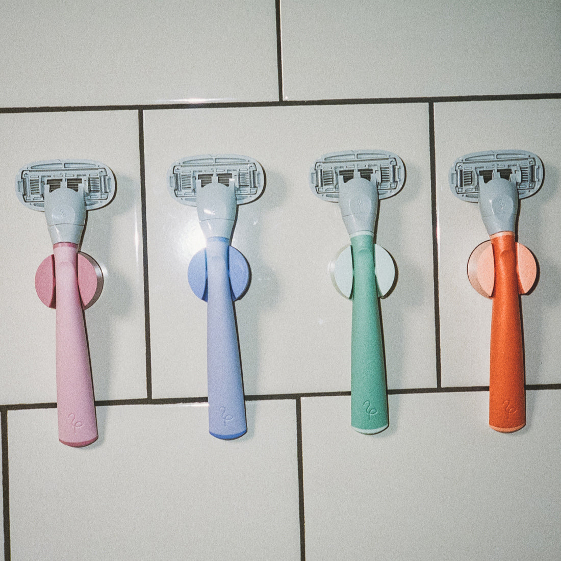 Rose, Lilac, Sage, and Papaya Flamingo Razors in their color coordinated shower holders on a tile bathroom wall