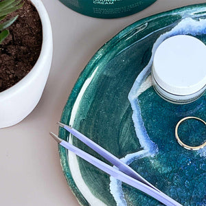 Flamingo Tweezers on a glazed teal ceramic dish that also holds a gold ring and a small jar of cosmetics 